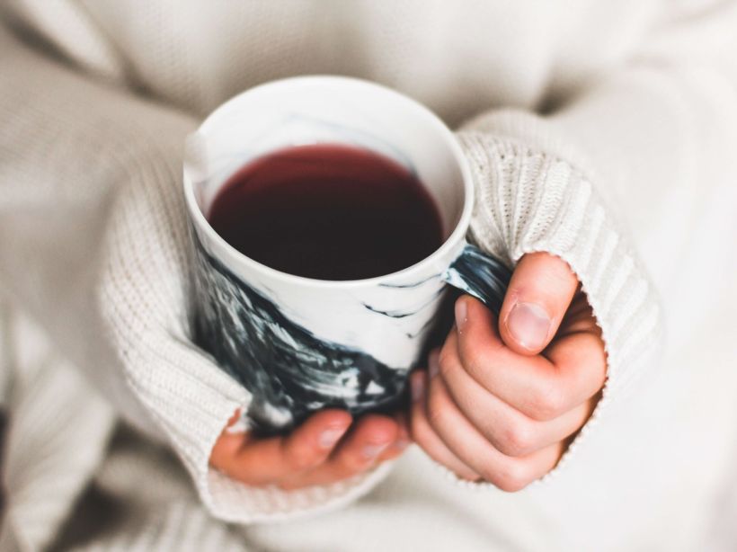 A close up of a person's hands, who is dressed in a cozy white sweater, holding a mug of hot black tea.