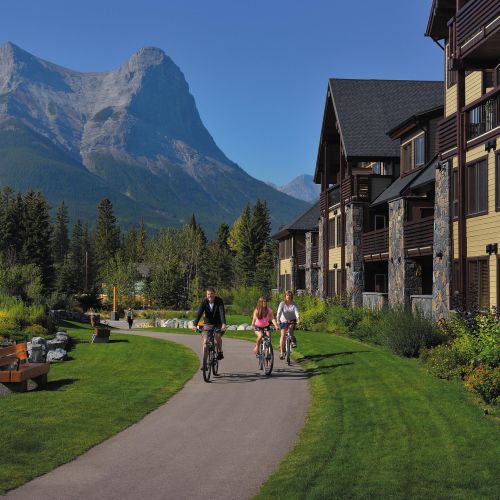 A group of people on bicycles cycle along a path lined with bright green grass and plants, with buildings on the right hand side, and mountains in the background in Spring Creek, Canmore, a more sustainable way to travel for an eco-friendly vacation.