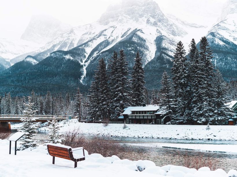 A wintery view of the Bow River by the Canmore Engine Bridge, with the snowy mountains and forests in the background, and fresh snow on the ground.
