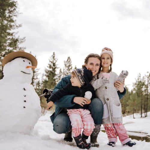 A mother crouches outdoors while hugging her son and daughter beside the snowman they built on a snowy winter day. Build snowmen with your family while on a family Christmas vacation in Canmore.