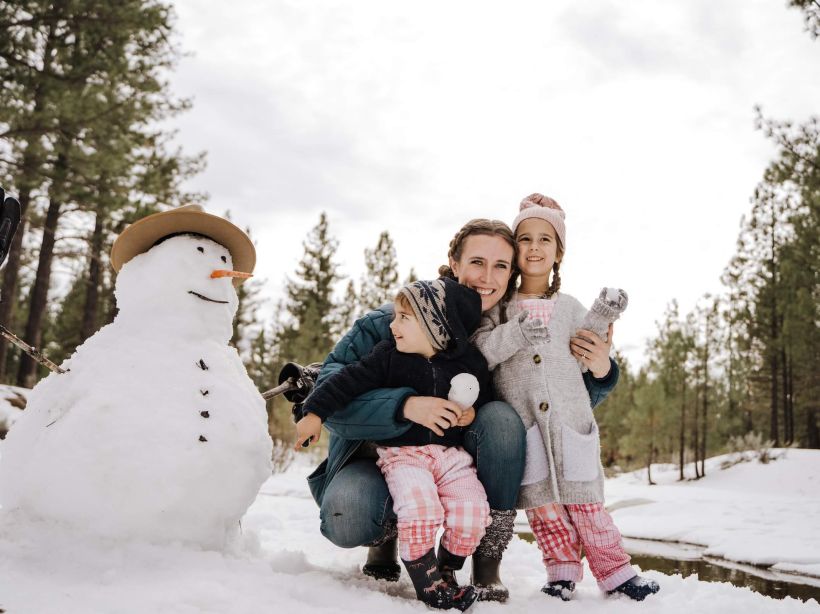 A mother crouches outdoors while hugging her son and daughter beside the snowman they built on a snowy winter day. Build snowmen with your family while on a family Christmas vacation in Canmore.