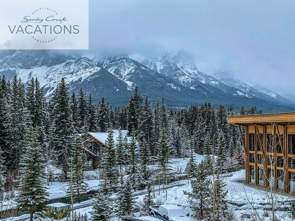 A building stands on the right, while snowcovered trees stand on the left, with mountains in the background and a low snow fog above in Canmore, Alberta at Spring Creek Vacations
