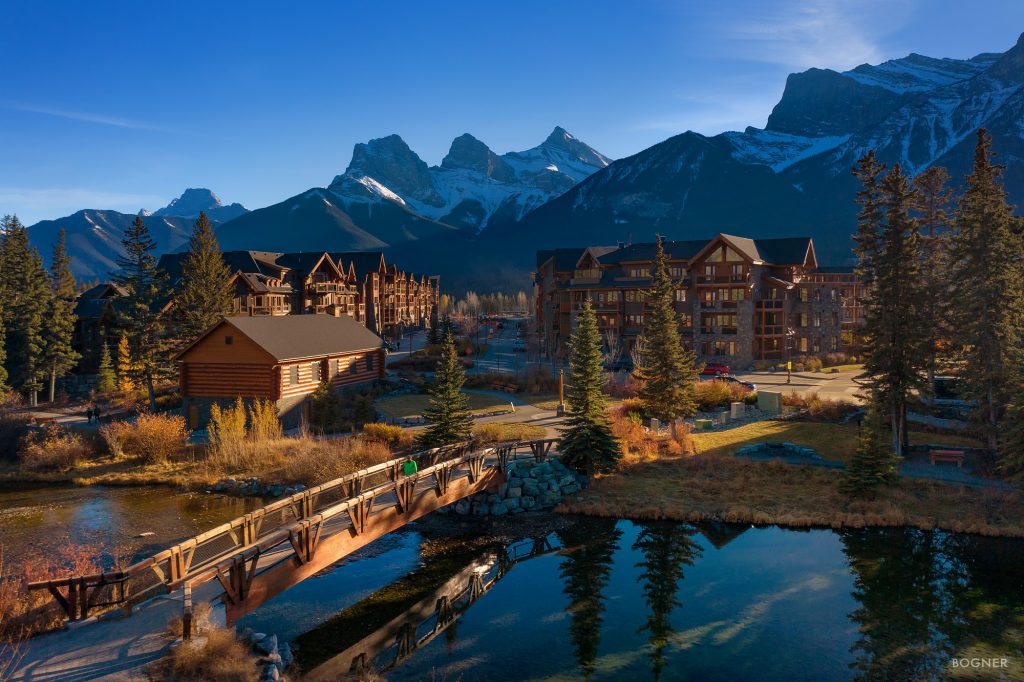 The Built Green certified Spring Creek community in Canmore, with the sun setting over the Canadian Rocky mountains and sustainably built buildings sprawling in the background, with a bridge arching over a creek.