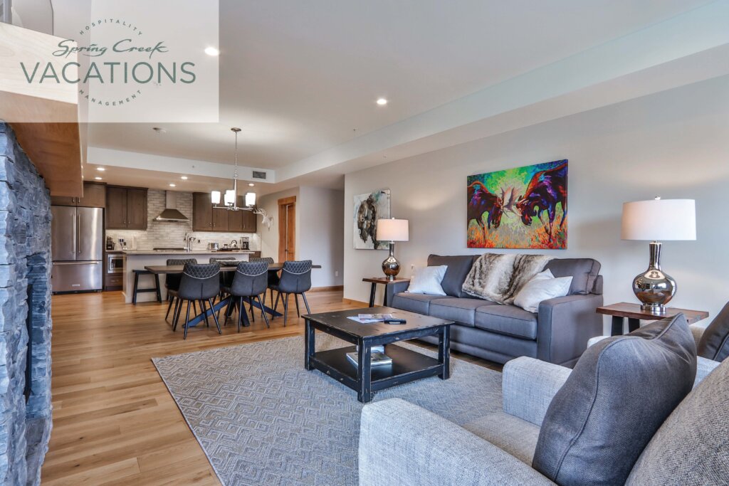 An open-concept living room and gourmet, luxury kitchen, with wooden floors, cream coloured walls, cozy grey couches and carpet, and modern decorations in a Spring Creek Vacations rental suite, in Canmore, Alberta.