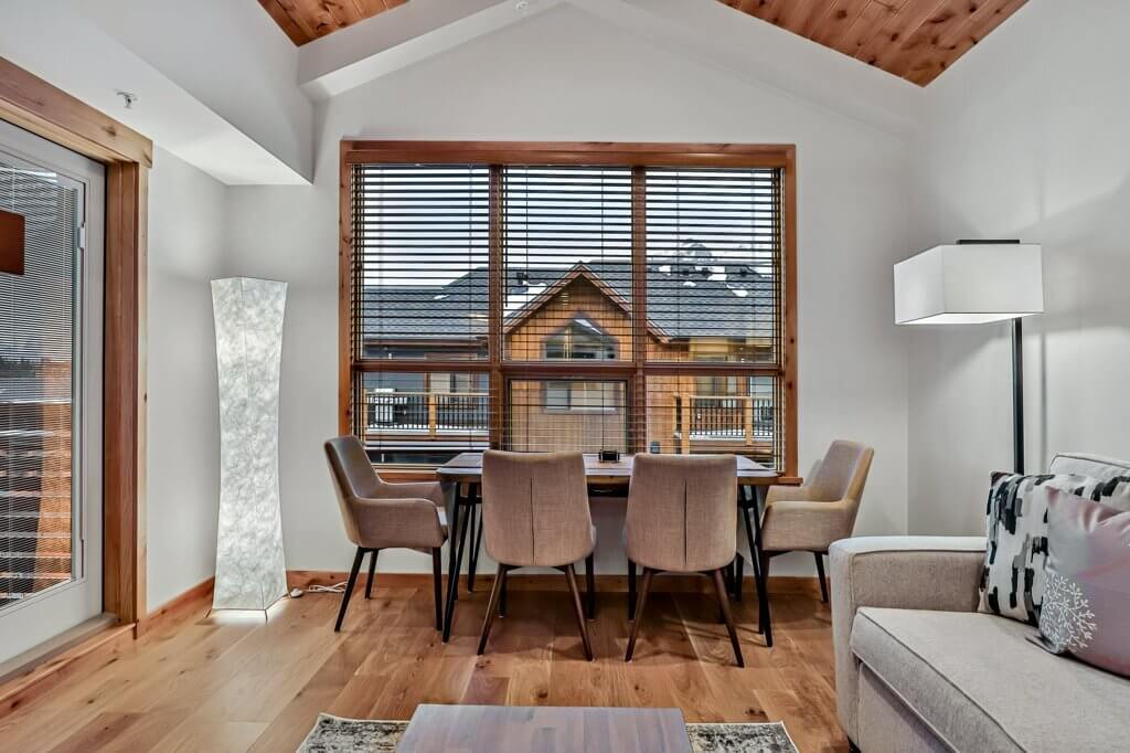 A living room and dining area in a Spring Creek Vacations luxury suite, with a large window overlooking another home in Canmore, Alberta, which has cream walls, wooden floors and a wood panelled roof, and modern lighting and decorations.