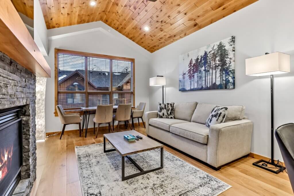 A stunning luxury living room and seating area with modern decorations that include a light grey couch, wooden coffee table, grey and blue carpet, and a wooden floor and wood panelled ceiling, in a Spring Creek Vacations suite in Canmore, Alberta.