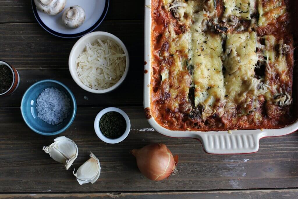 a bird's eye view shot of a baked lasagna in a ceramic dish, with ramekins of shredded cheese, salt, and herbs nearby, as well as an onion, mushrooms, and garlic.