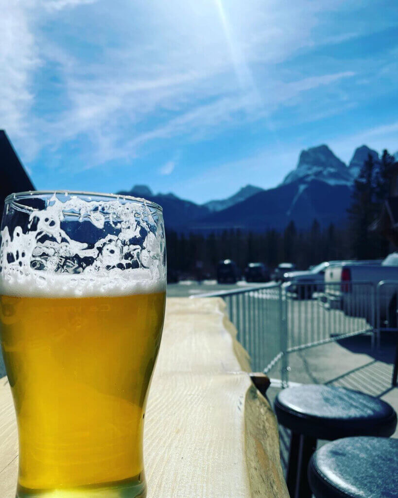 A light golden beer in a beer glass, partially drunk, placed on a wooden counter, in close focus, with Canmore's mountains in the background, from Sheep Dog Brewing in Canmore, Alberta.
