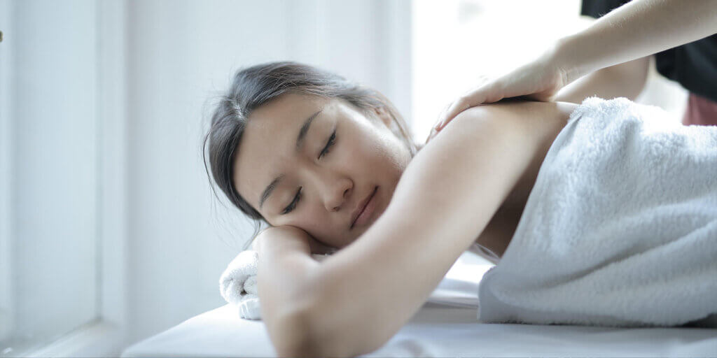 A woman with her eyes closed and in a white towel smiles as she is lying down getting a back massage in a white room, in Canmore, Alberta.