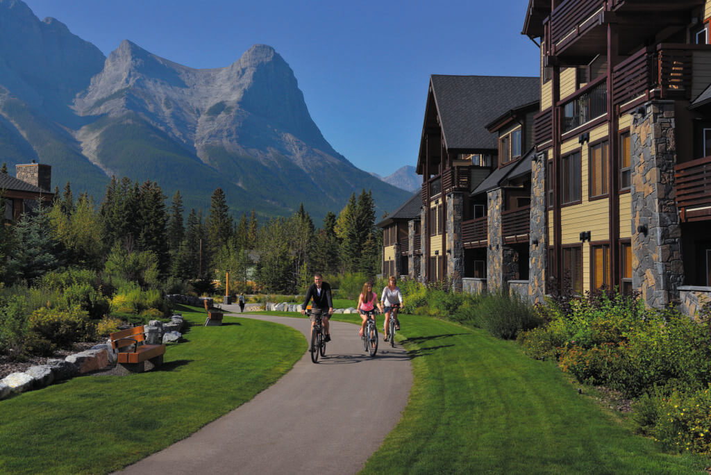 A group of people on bicycles cycle along a path lined with bright green grass and plants, with buildings on the right hand side, and mountains in the background in Spring Creek, Canmore, a more sustainable way to travel for an eco-friendly vacation.