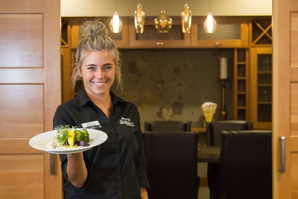 Female server in black uniform stands with a plate of food in her right hand, smiling while at Mineshaft Tavern in Spring Creek Mountain Village