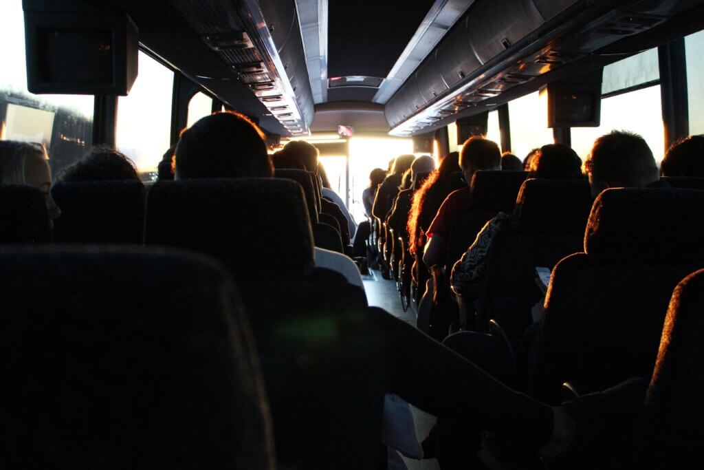 A shadowed view of the inside of a travel bus, packed with tourists and travellers in every seat on a sunny day.