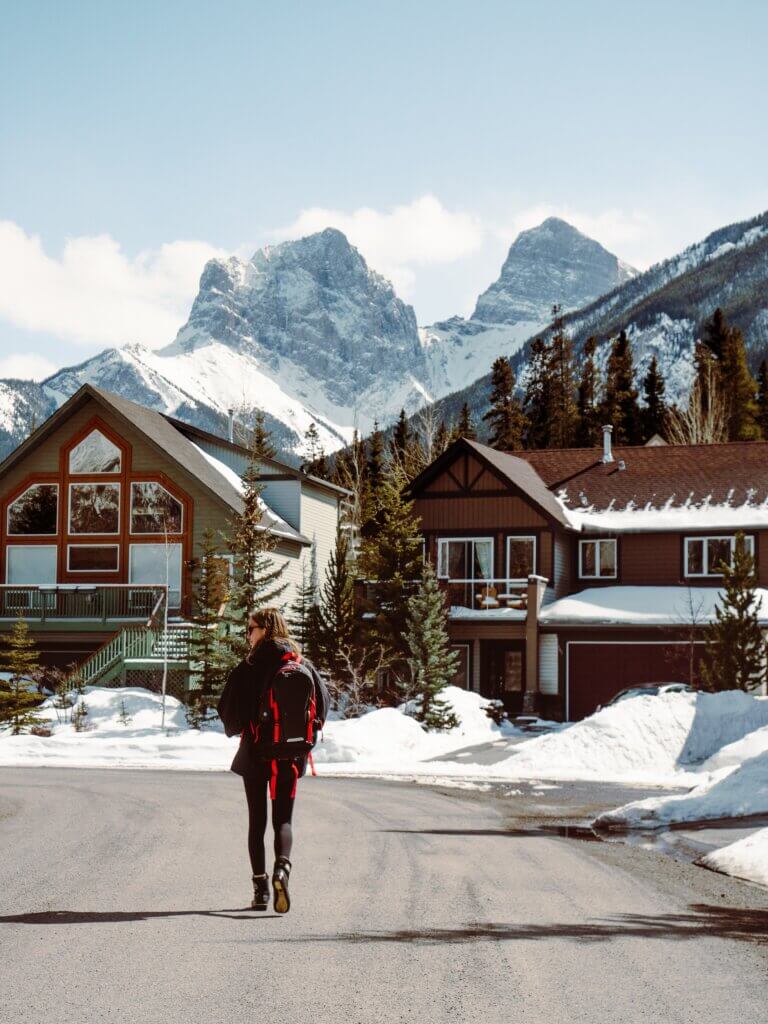 A woman dressed in black with a red backpack walks through a road in snowy Canmore, Alberta, surrounded by brown buildings and homes covered in snow, with mountains in the background, on a sunny day.