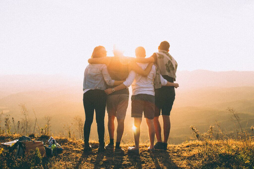 Four people stand with their arms around one another at the top of a mountain, looking our towards a seeting sun during golden hour, while on a group trip.