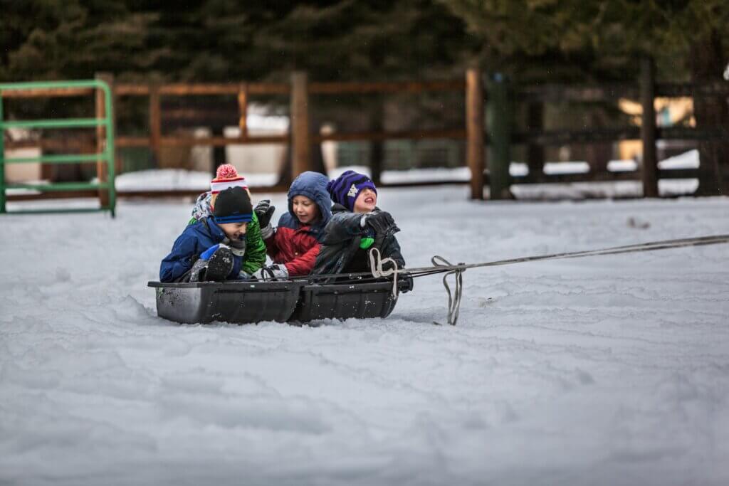 Four kids ride on a toboggan getting pulled through the snow with trees and a wooden fence in the background, having some winter Christmas holiday fun