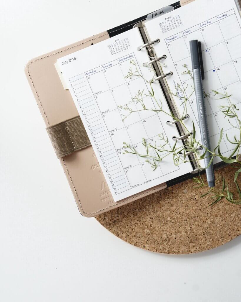 A day planner with a light beige leather cover and spiral binding lies flat and open on top of a corkboard mat and table, with a pen laying across the paper as well as a sprig of greenery.