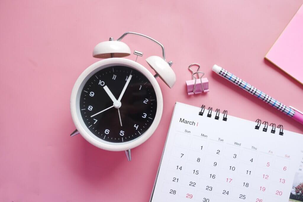A ringer-style mechanical clock lies on its back on top of a pink background, with a calendar, clip and pen beside it.