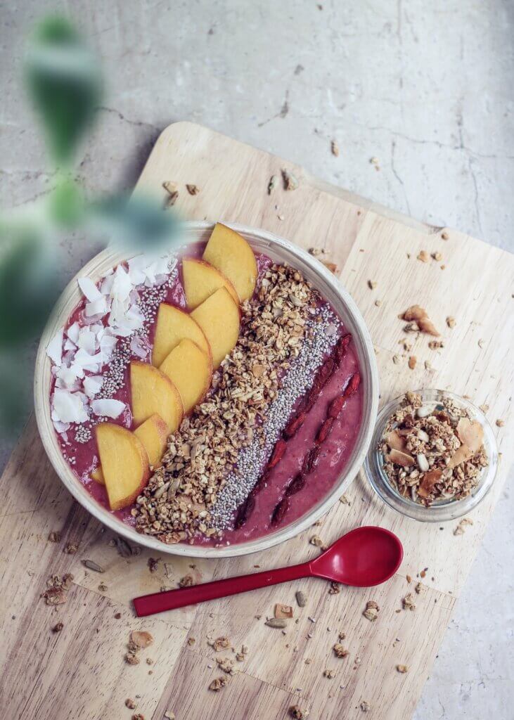 smoothie bowl topped with coconut, peach, granola and chia seeds with a red spoon beside on a wooden board