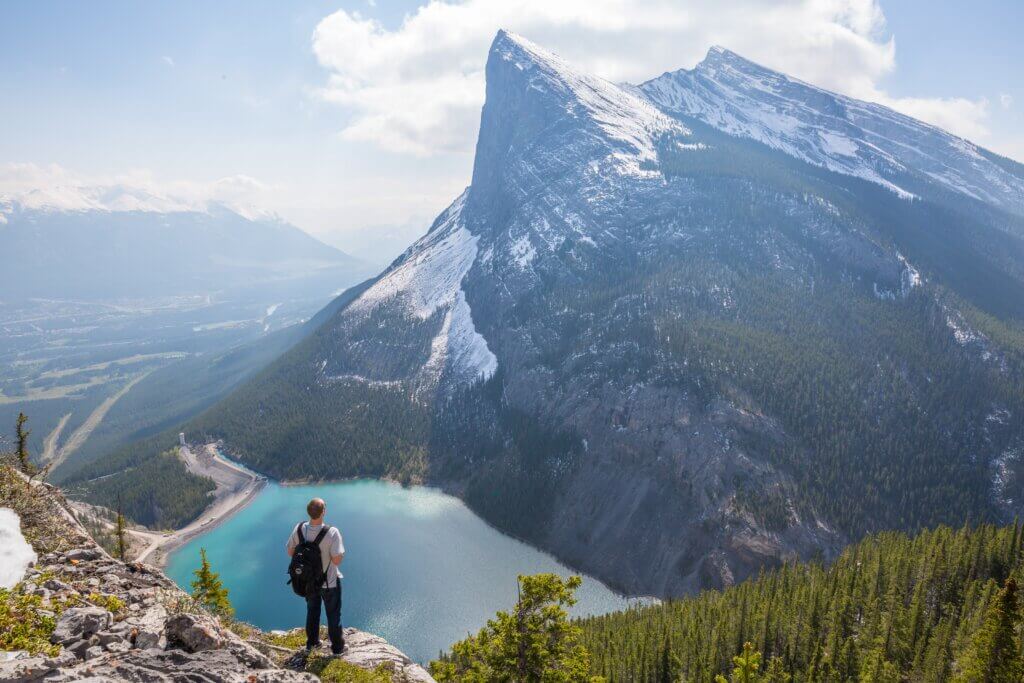 man standing on a cliff looking at the rocky mountains and lake below