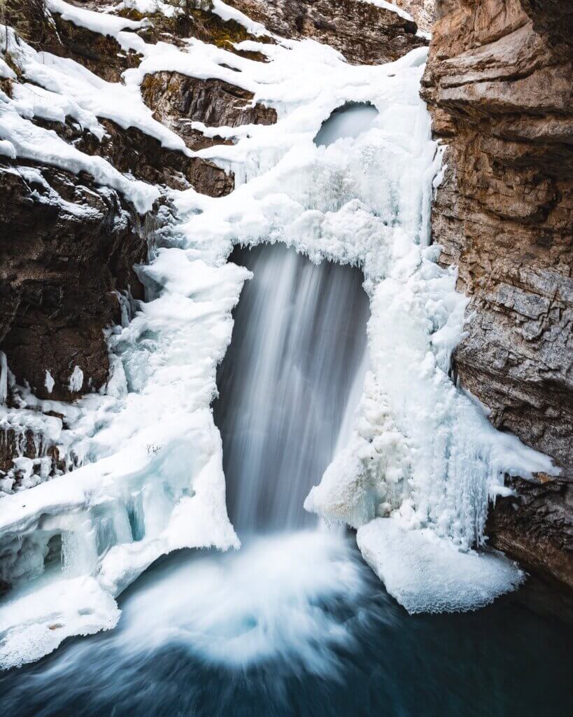 A dramatic, frozen waterfall with giant rock formations surrounding it, as two hikers travel beside the formation in the distance, at Johnston Canyon near Canmore, taken in Canmore during the winter.