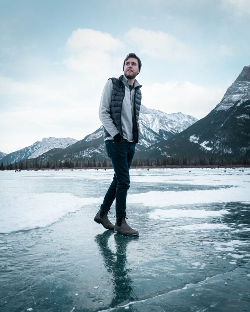 A man in a grey sweater and dark vest with a toque stands on a frozen, reflective lake with patches of snow around him and Canmore's mountains in the background while doing a winter hike in Canmore, Alberta.