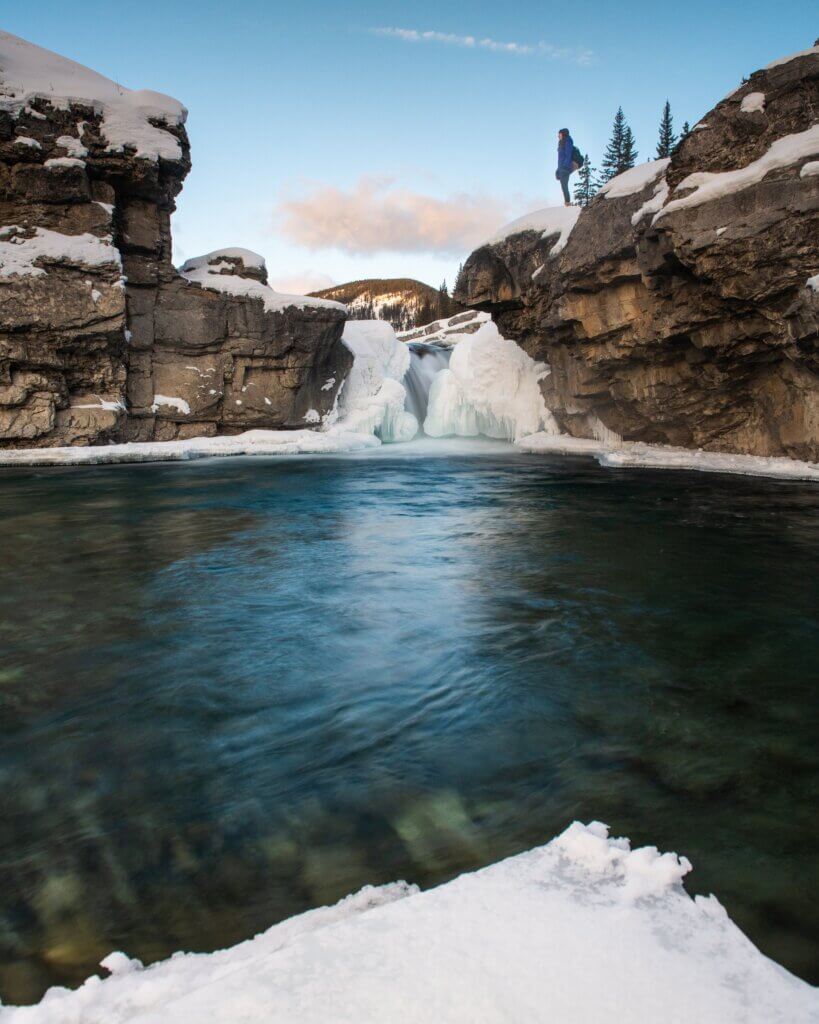A crystal clear lake surrounded by snow and dramatic rock formations stretches out, with a frozen waterfall in the background and bright blue skies, while a woman in a blue jacket stands near the edge of the rock formations, taken in Canmore during the winter.