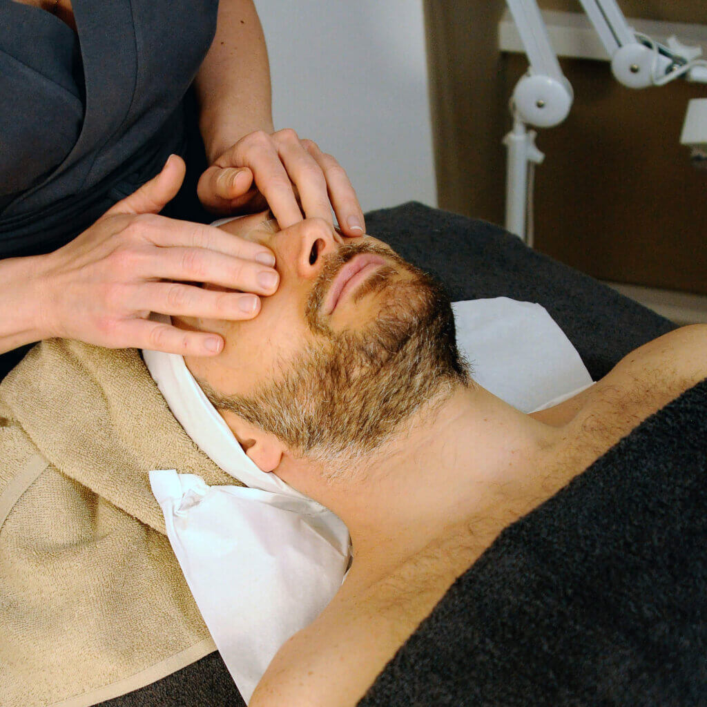 A bearded man relaxes and lays face up while a masseuse massages his face.