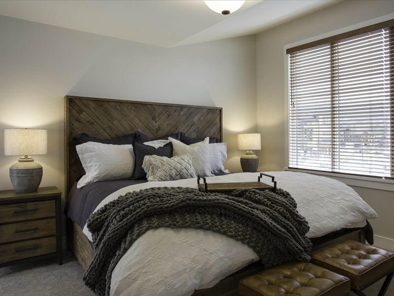 A luxurious but cozy bedroom with a comfortable bed, neutral toned colouring, bright light fro a large window and wooden accents in a Tamarack Lodge luxury condo.