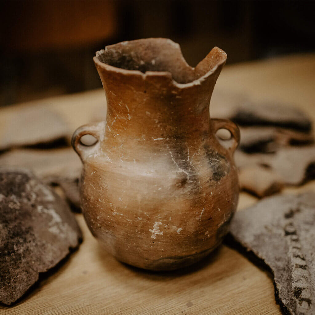 An ancient artefact style vase lays with other broken pieces of artefacts on a wooden table.