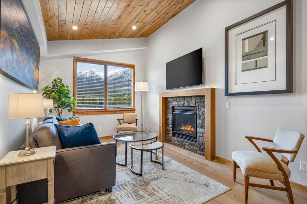 Tamarack Unit 403A's living room with a cozy stone fireplace, bright natural light, hardwood floors, and modern finishes located at Spring Creek Vacations in Canmore, Alberta.