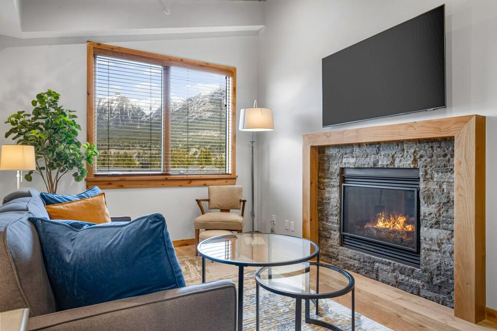 Tamarack Unit 403A's living room with a cozy stone fireplace, bright natural light, hardwood floors, and modern finishes located at Spring Creek Vacations in Canmore, Alberta.