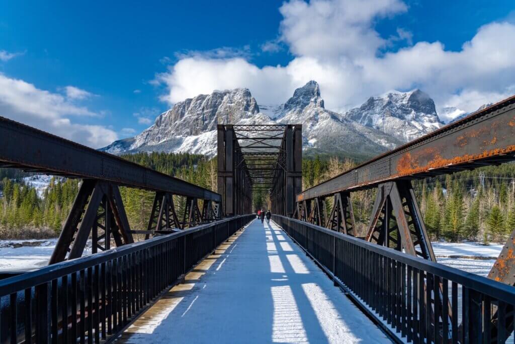 A shot from one end of the Canmore Engine Bridge to the other, on a sunny day in winter with snow around, with mountain peaks and blue skies in the background, one of the best photo spots in Canmore.