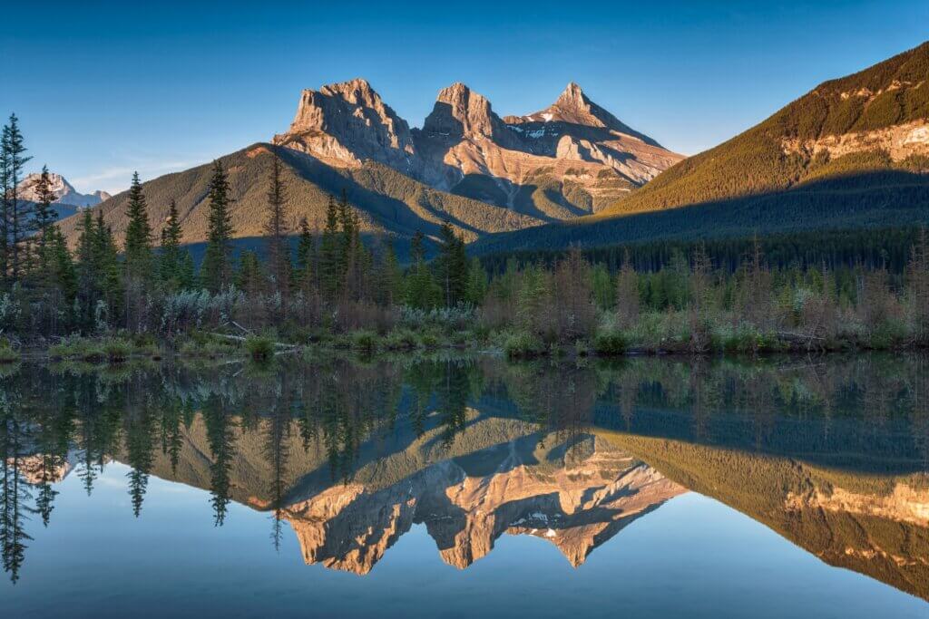 A view of the Three Sisters Peaks from Policeman's Creek Viewpoint in summer, with bright blue skies, a golden sun shining on the mountains, and still, reflective waters ahead, one of the best photo spots in Canmore.