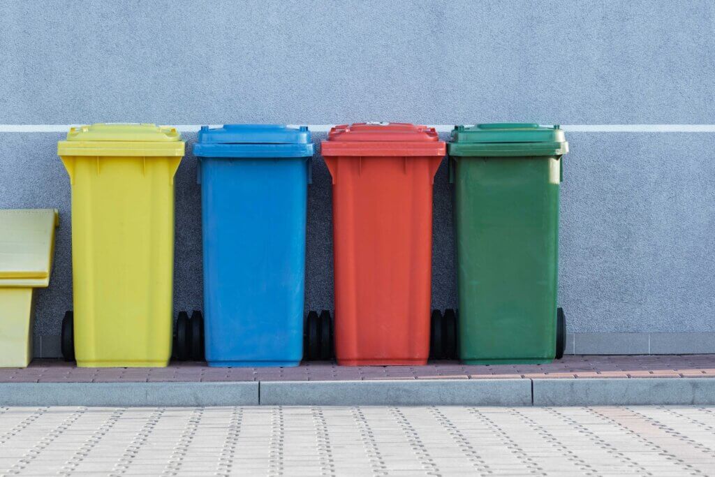 Four bins for recycling and disposing of waste, with one yellow, then one blue, then a red, and finally a green coloured one, stand against the wall of a building on a sunny day.