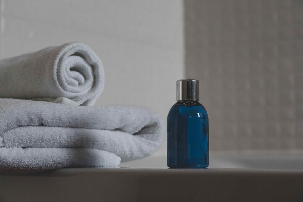 A reusable travel-sized bottle of blue shampoo sits ont he edge of a bath next to some white towels in a bathroom, perfect to use for an eco-friendly vacation.