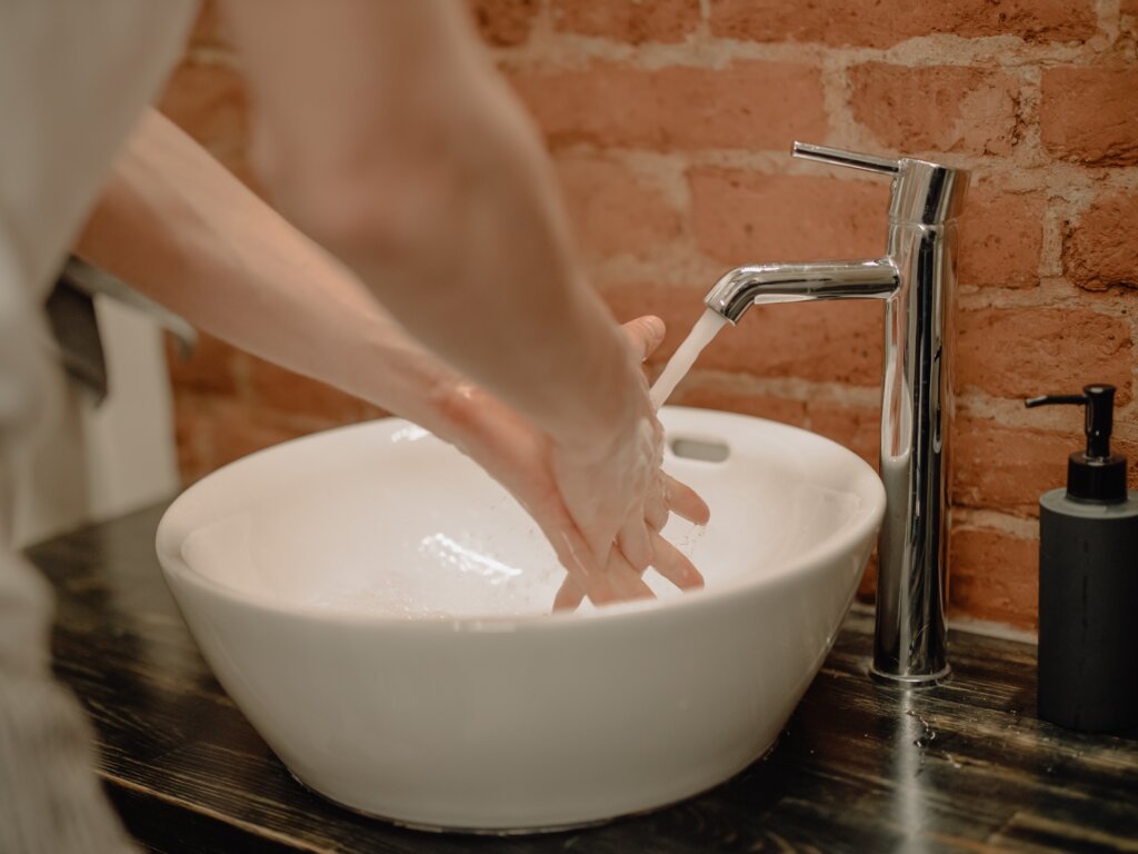 A man washes his hands in a white bowl sink with the water running in the bathroom, which has a brick wall and a wooden countertop.
