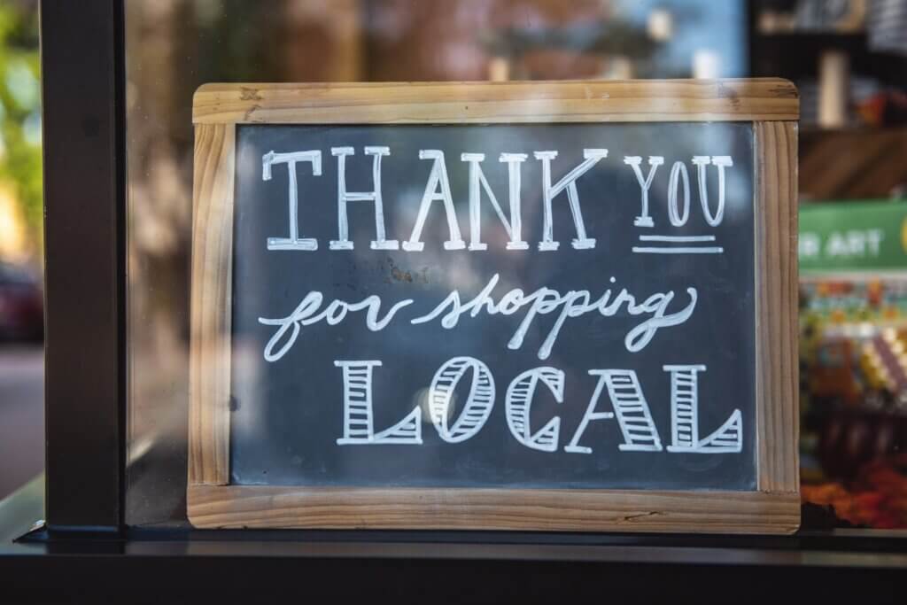 A chalkboard sign with a wooden frame that reads thank you for shopping local sits in a glass window of a shop.