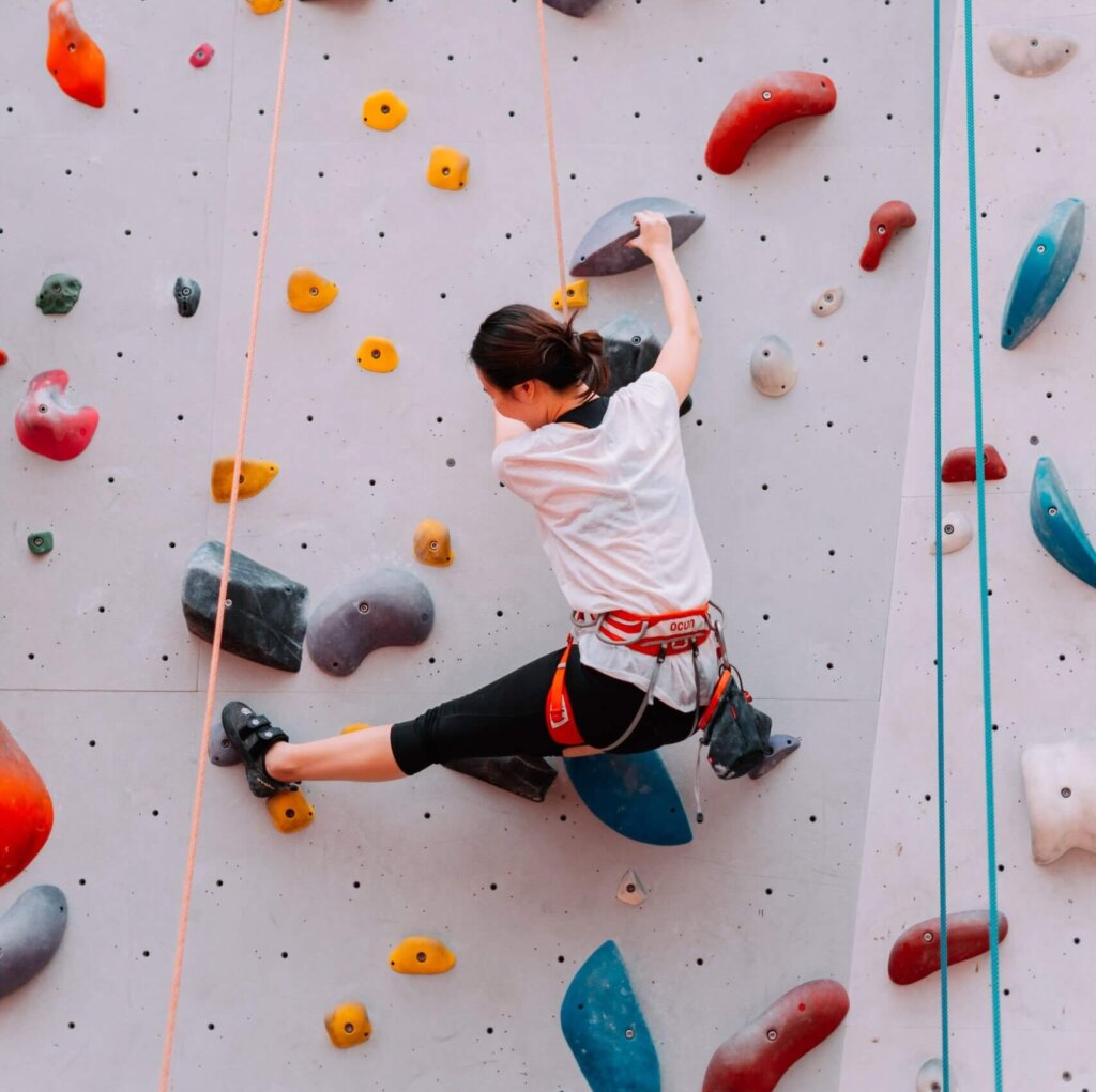 A woman in active wear and in a harness climbs an indoor rock climbing wall.