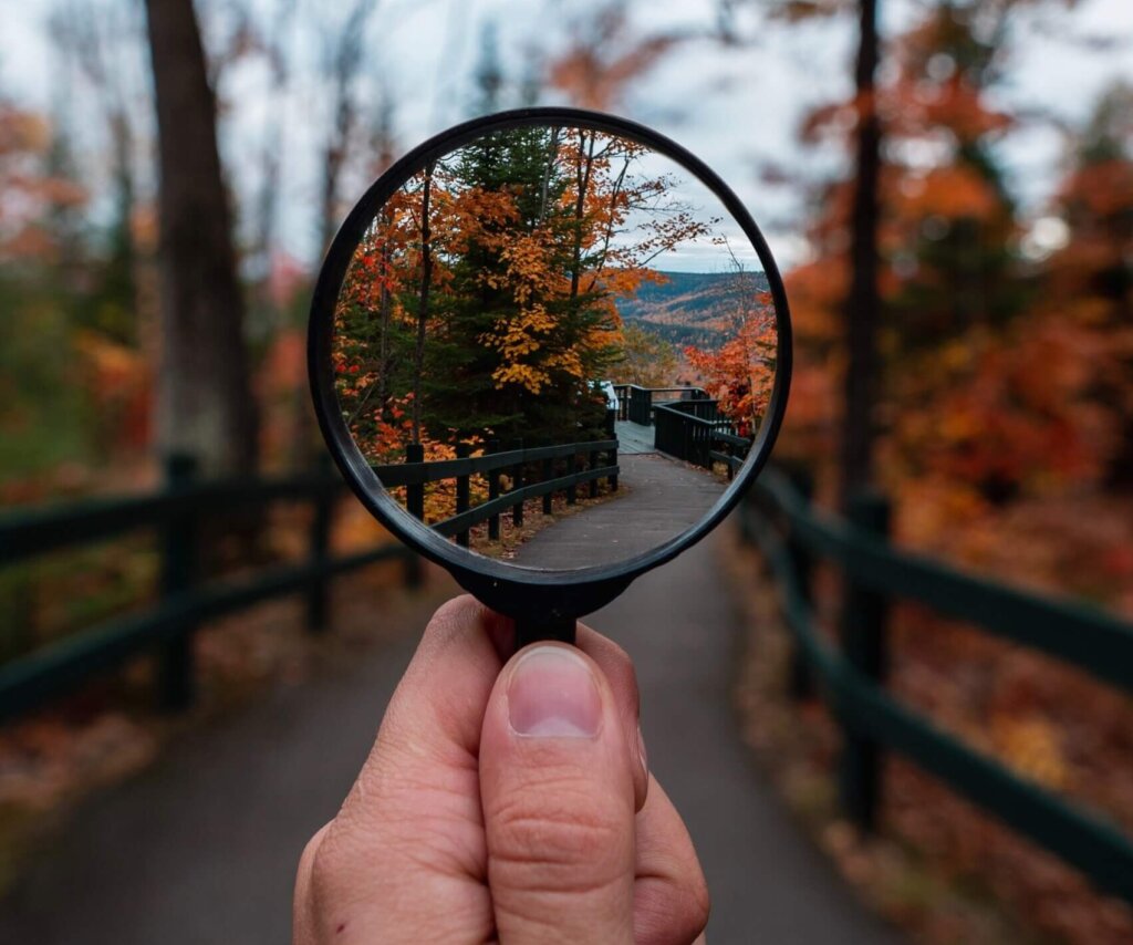 A man's hand holds a black rimmed magnifying glass while looking up at a fenced path between trees turning gold in the fall season.