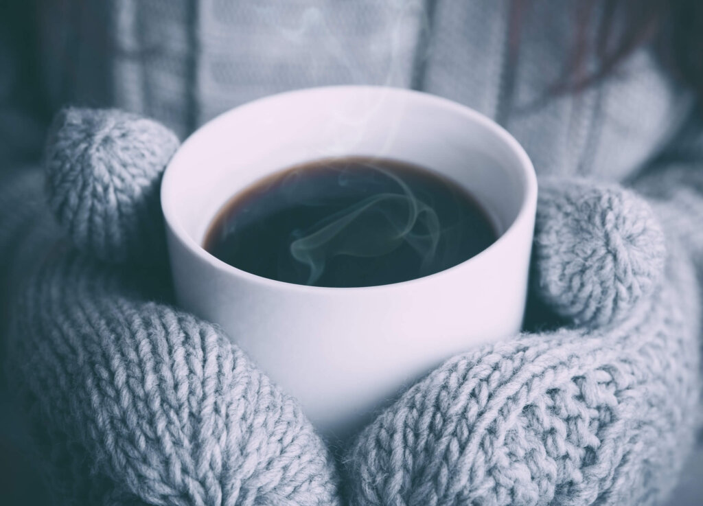 Two hands with knit mittens in a light grey colour hold a white ceramic mug with hot coffee, a perfect winter treat to get cozy with.