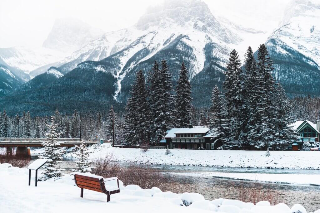 A wintery view of the Bow River by the Canmore Engine Bridge, with the snowy mountains and forests in the background, and fresh snow on the ground.
