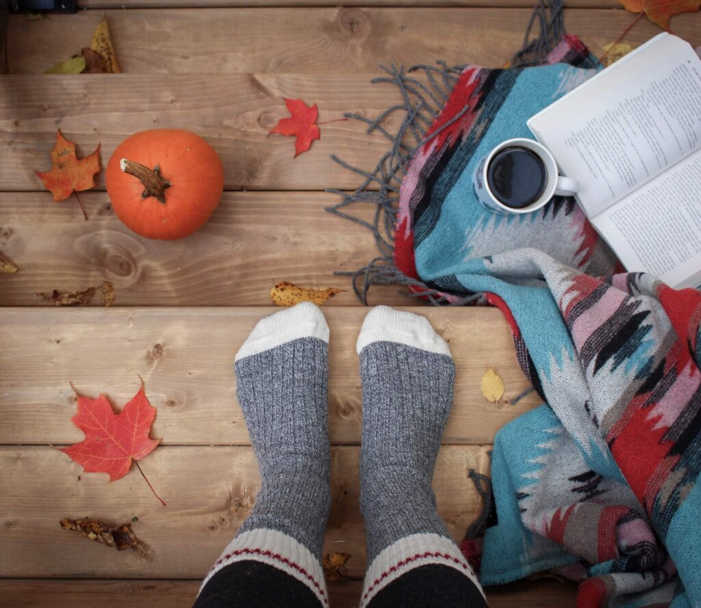 A pair of feet with warm socks stands on wooden steps with a blanket, book and hot drink on the right, and a mini pumpkin on the left.