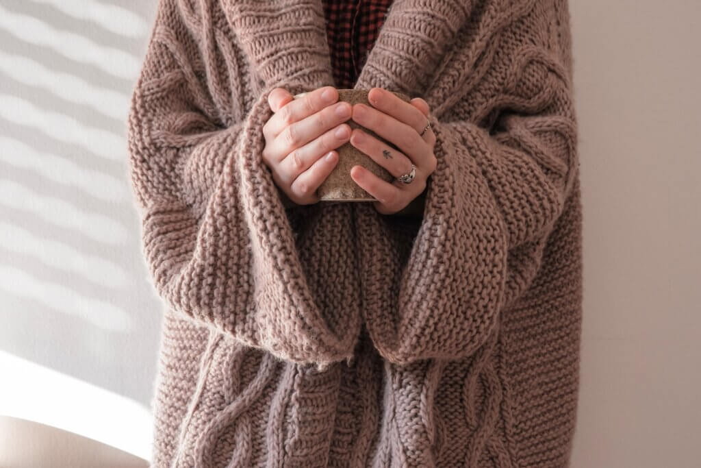 A woman in a light brown warm knit sweater holds a mug of a hot drink in her hands to get cozy.