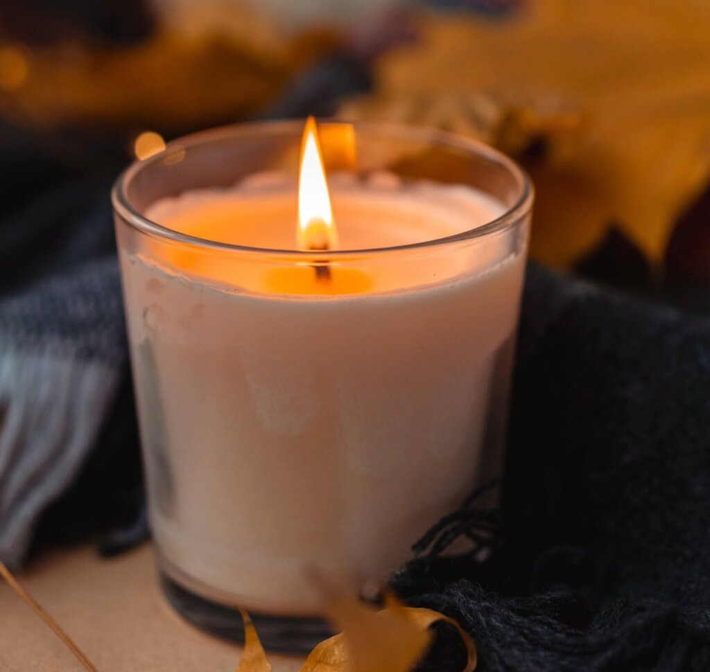 A lit candle in a glass container sits on a fall decorated table with a table cloth, fall leaves and a white pumpkin in the background.