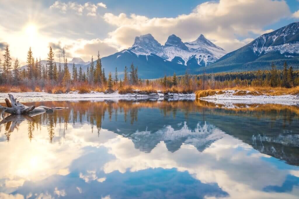 A view of the Three Sisters peaks over a lake in winter in Canmore, with snow dusted ground and bright sunlight sparkling off of the still water, a beautiful view in winter and a great activity to try in Canmore in winter.